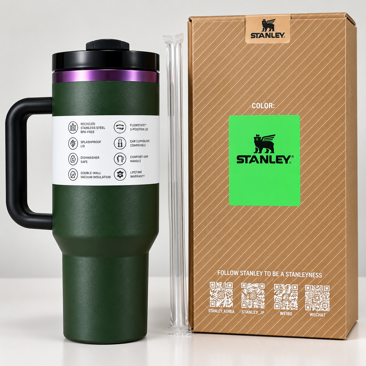 A dark green Stanley travel mug with a black handle and lid, shown next to its brown cardboard packaging box and a clear straw on a light gray background.