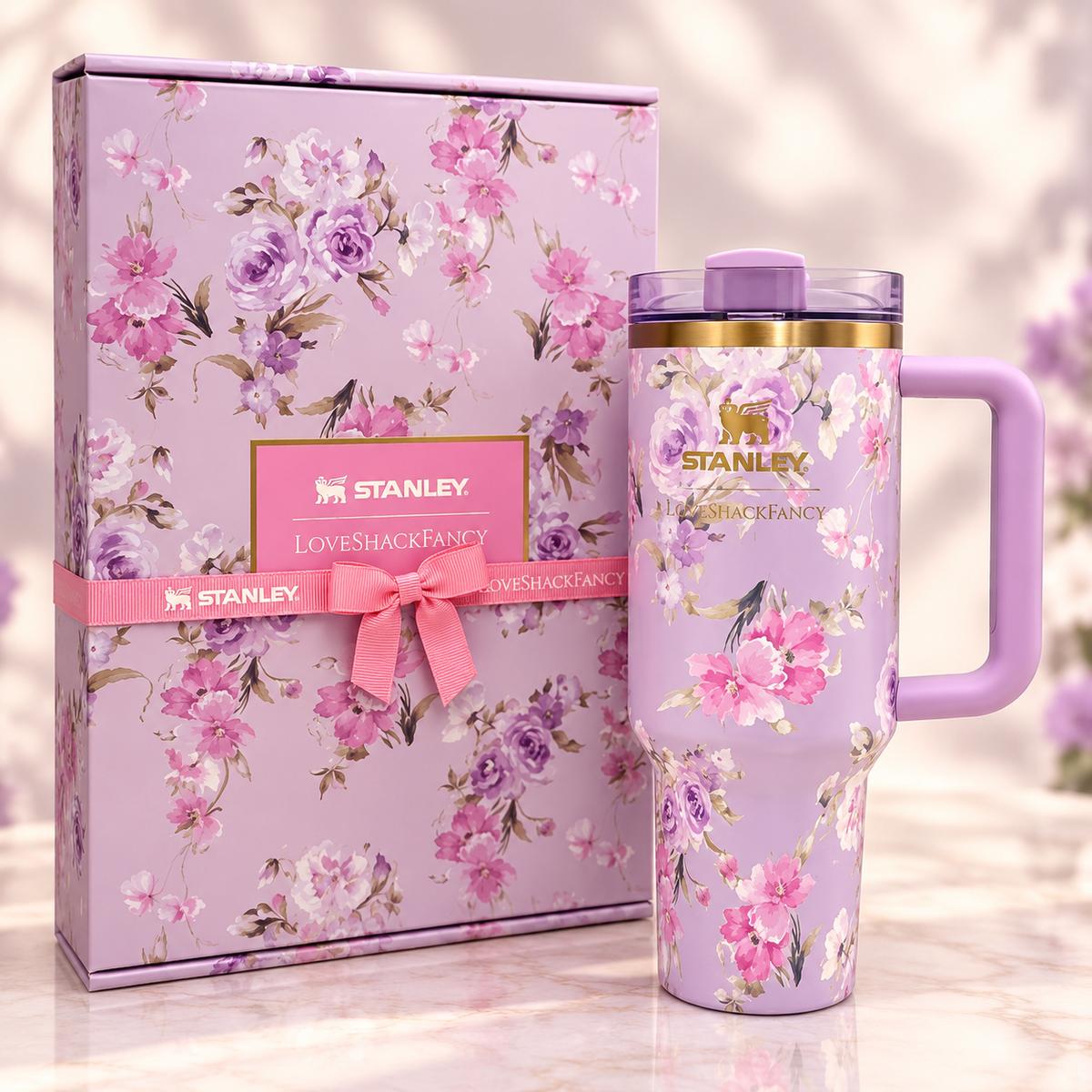 A lavender floral Stanley travel tumbler with a purple handle and lid, shown next to its matching floral gift box with a pink ribbon and visible Stanley branding.
