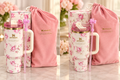 A Stanley tumbler with a pink floral print, shown alongside its pink drawstring bag and matching box on a light marble surface. The tumbler has a white handle, a pink lid, and a pink straw with a bow, and the Stanley logo is visible on the bag and tumbler.