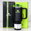 A black Stanley travel mug with a handle and clear lid, shown with its branded black spiderweb-pattern box, lime green packaging, and a clear straw on a light gray background.