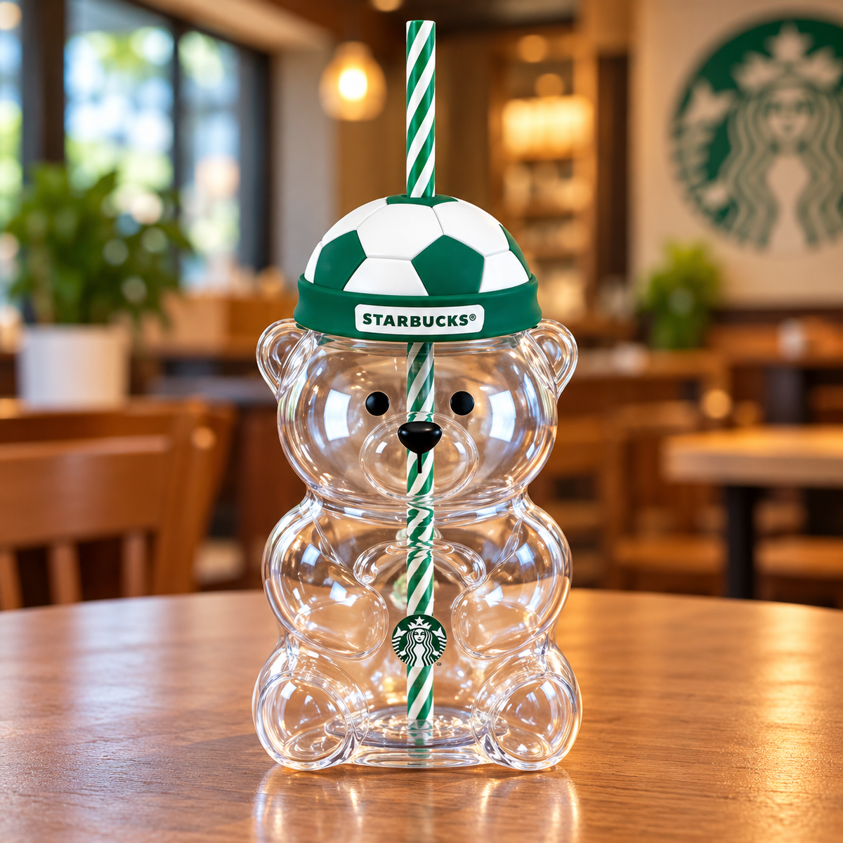 A clear bear-shaped Starbucks tumbler with a green and white striped straw and a green-and-white soccer-patterned lid. The lid has a visible Starbucks logo, and the tumbler is photographed on a wooden table in a softly blurred Starbucks interior.