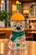 A clear bear-shaped Starbucks Peanuts tumbler with a green knit scarf, orange lid, and green-and-white striped straw, set on a wooden table in a warm, blurred cafe interior.